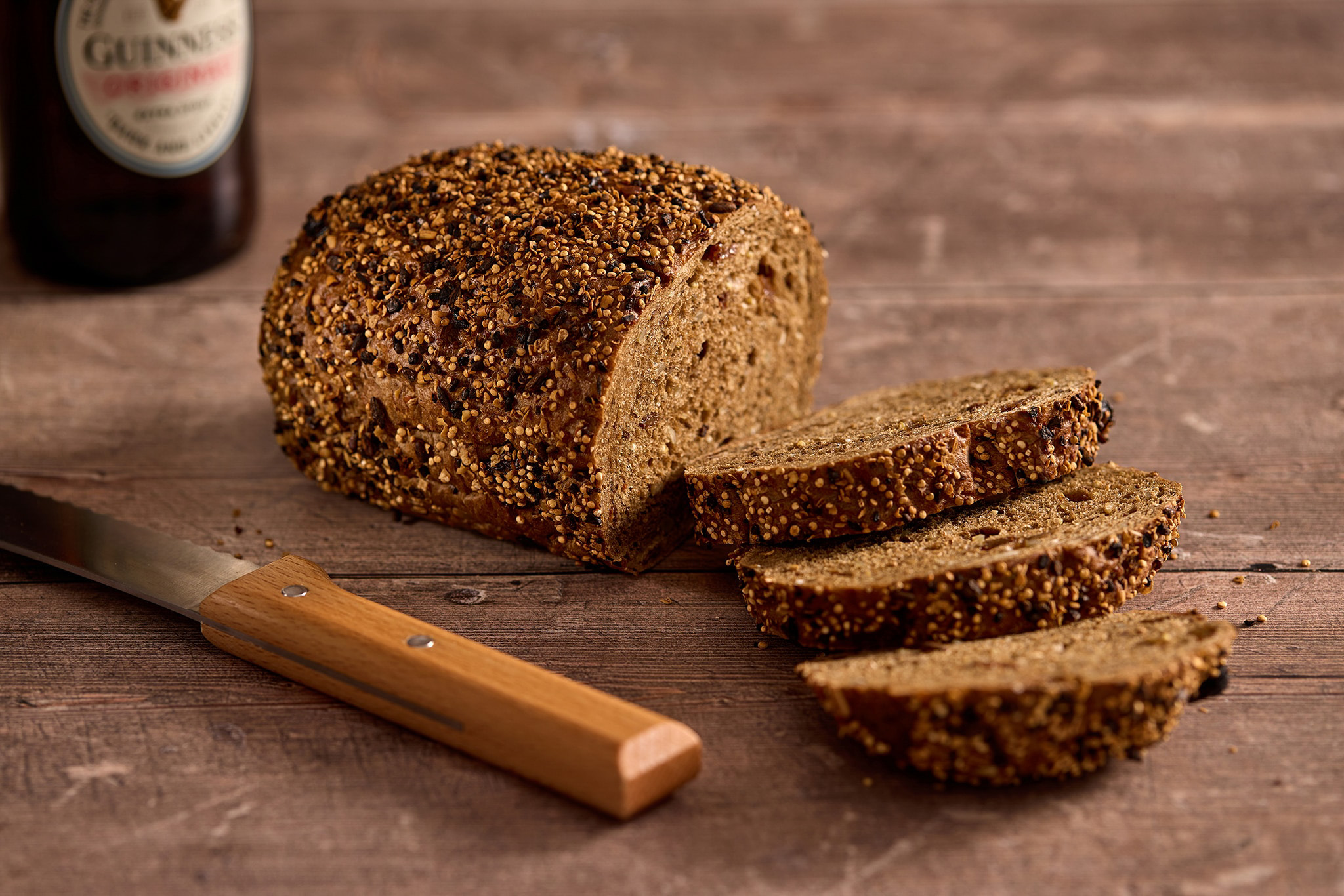 Dark brown seeded bread on a wooden board and a large bread knife next to it. Three slices have been taken out of the bread 