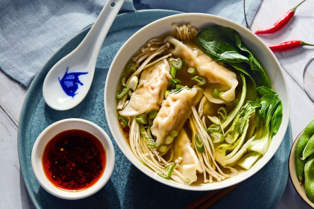 Bowl of chicken broth with asian greens and three gyoza. Next to it is a traditional chinese soup spoon and some chilli oil