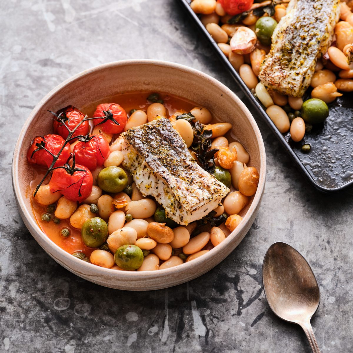 Cod dish with tomatoes and chickpeas roasted in the oven on a grey background with an antique spoon