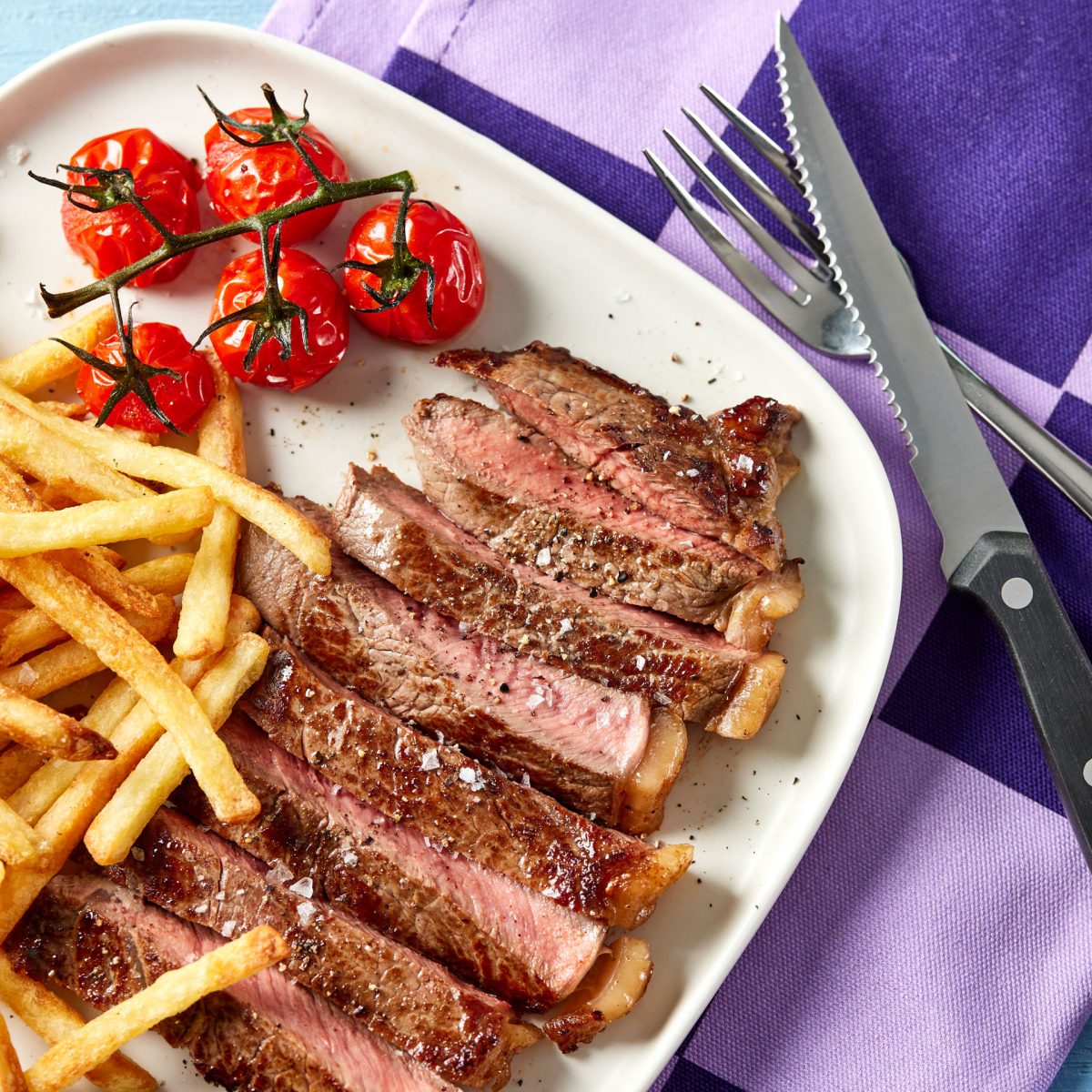 Sliced sirloin steak on a square plate with chips and roasted cherry tomatoes on a purple background