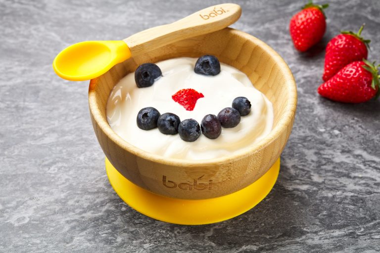 Yoghurt in a bamboo bowl with fruit used to make a face. Blueberries for the eyes and mouth and a raspberry nose