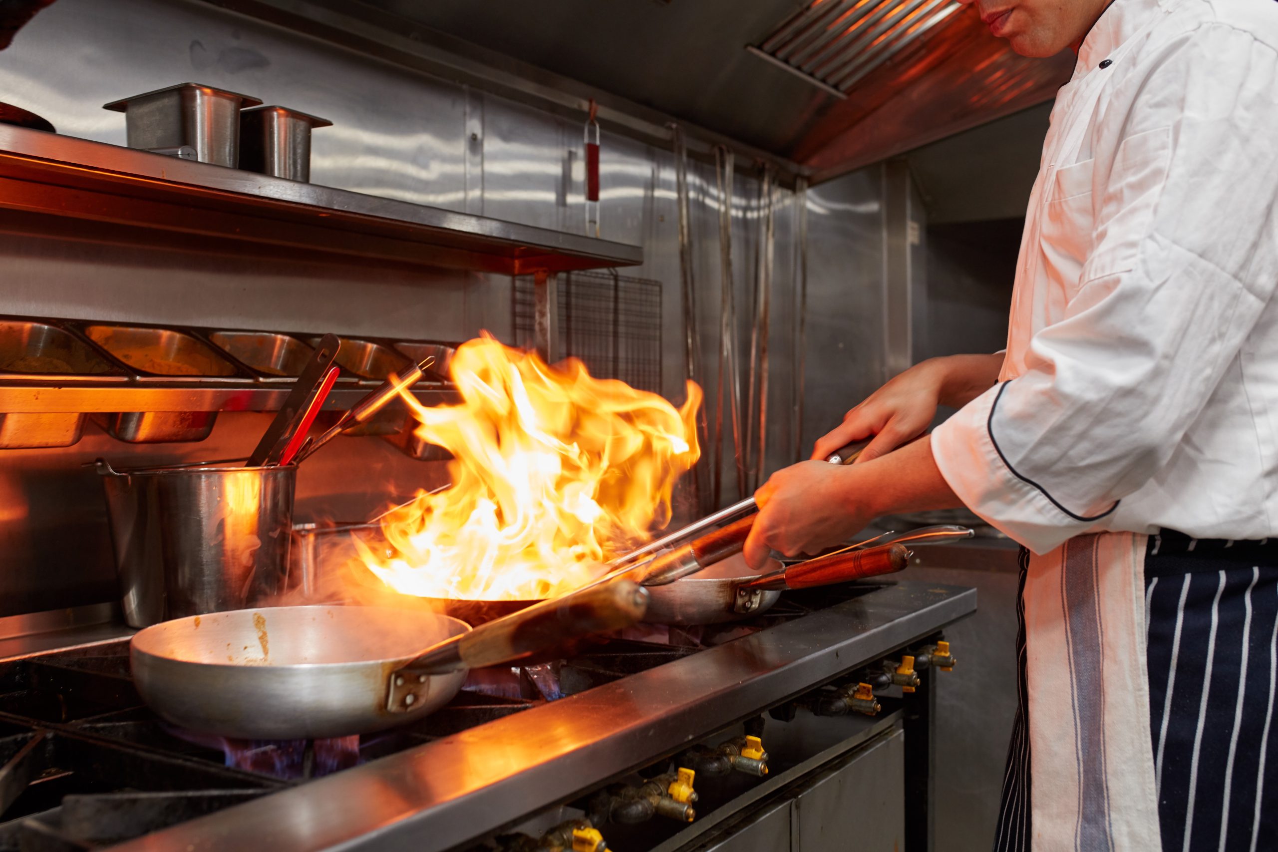 Chef executing flambé technique with fiery pan in kitchen.