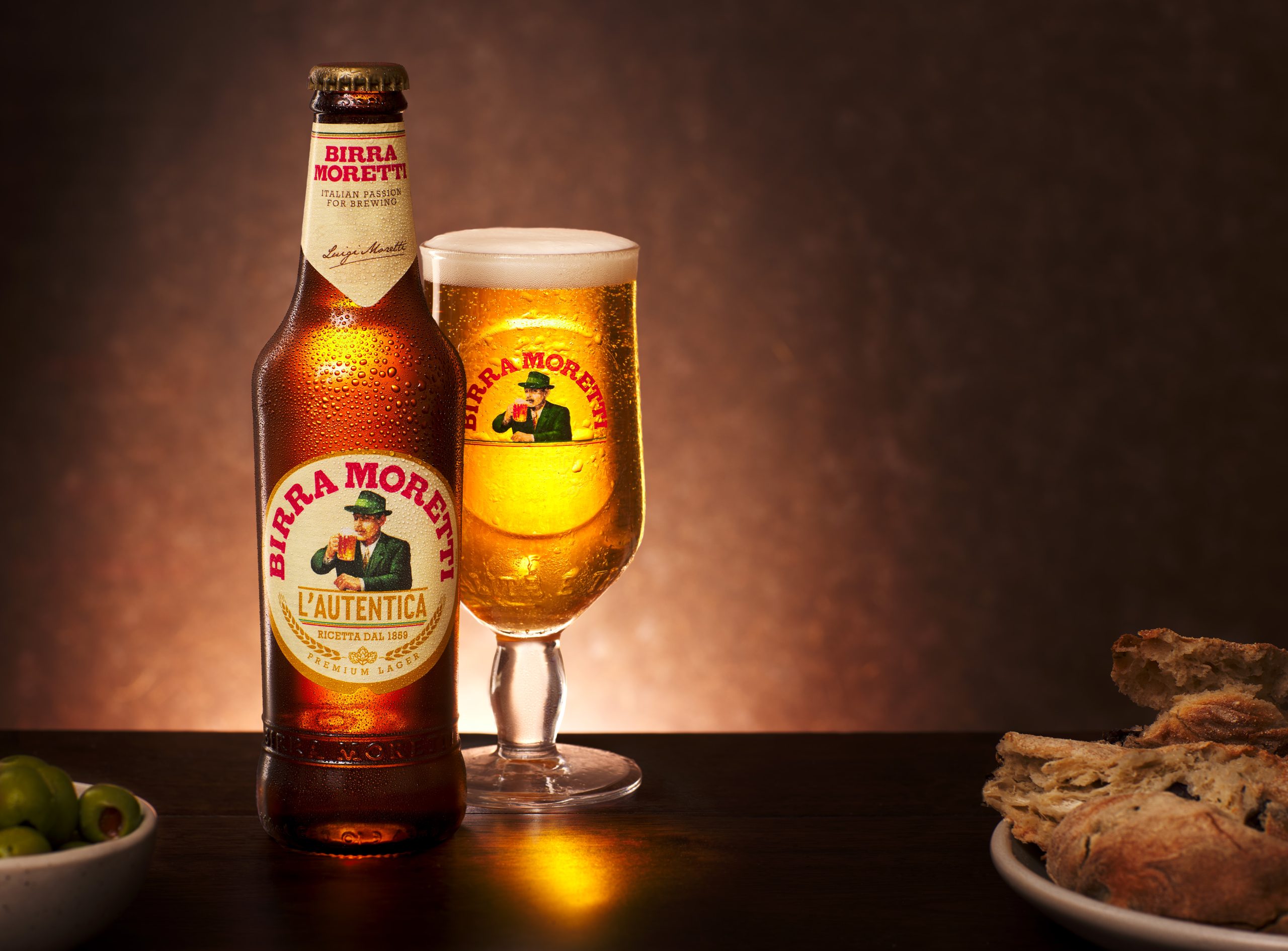 Birra Moretti with glass, olives, and bread on a warm-lit wooden table.