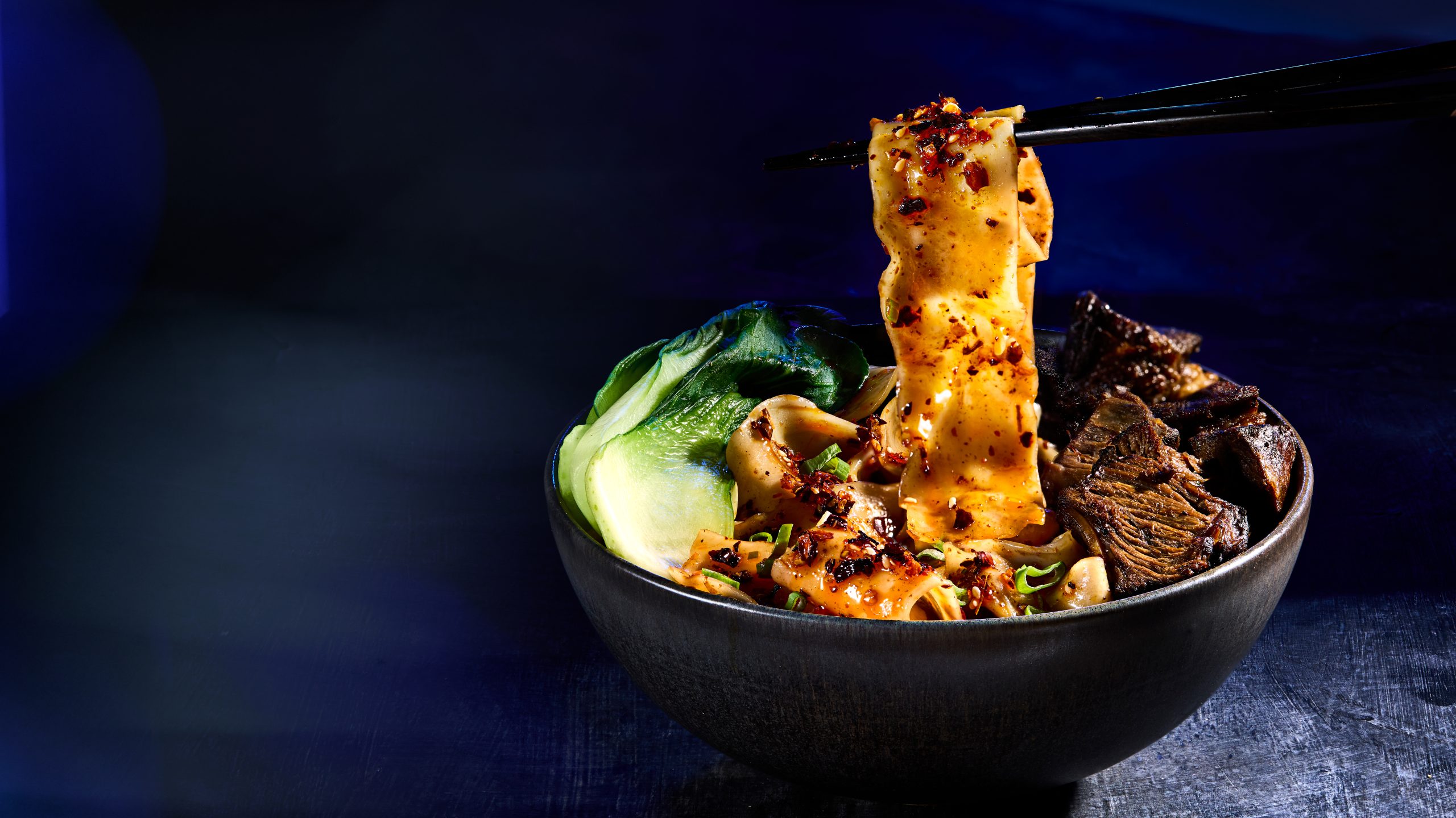 Spicy wide noodles with braised beef and bok choy lifted by chopsticks in a dark bowl.